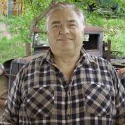 Large silver-haired man wearing a plaid shirt and standing in front of a rusted antique vehicle